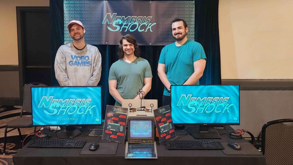 The Developers of Nemesis Shock standing in front of a banner and behind two computer monitors that read 'Nemesis Shock' at Seattle Indies Expo